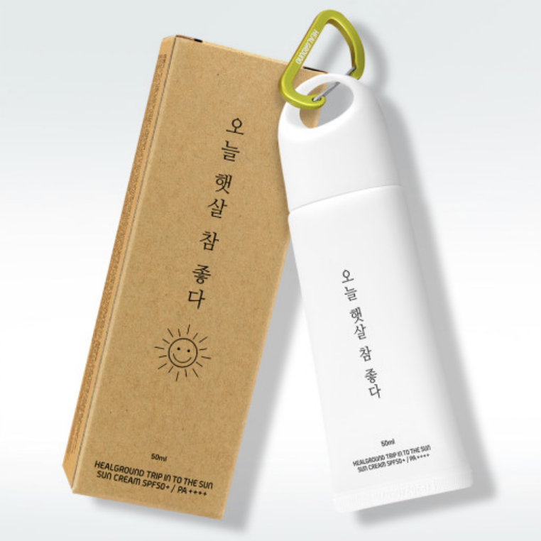 A slim white suncream tube with a green cap and a cardboard box with Korean text and a sun design. The box has a carabiner clip attached to it.